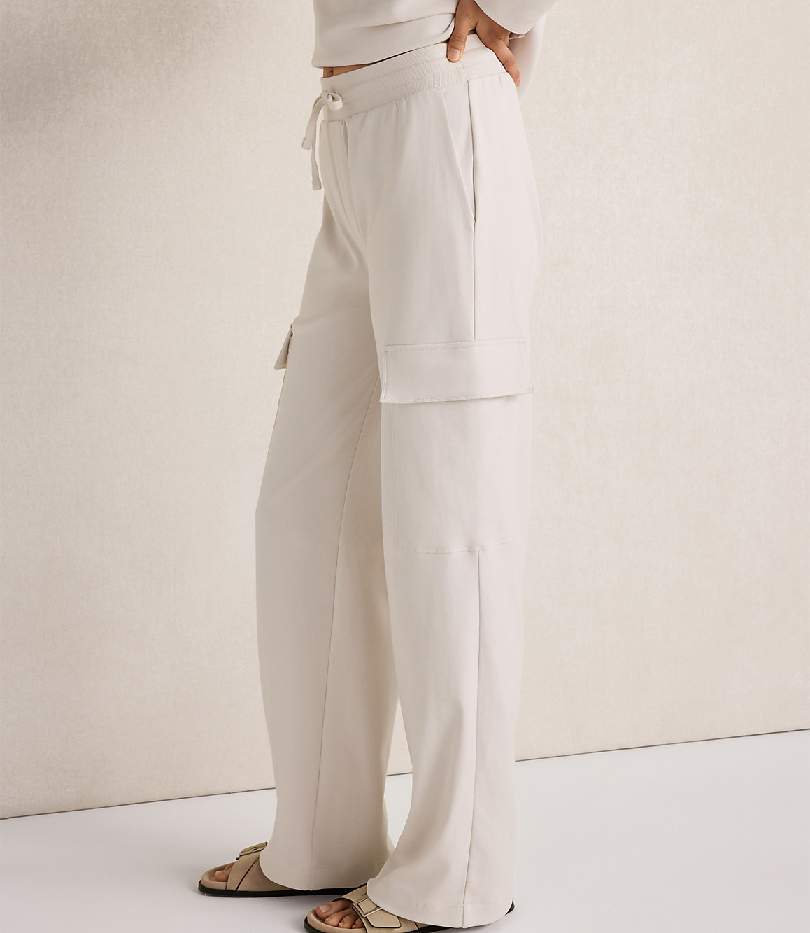 A person wearing cream-colored cargo pants and sandals stands against a textured cream wall.
