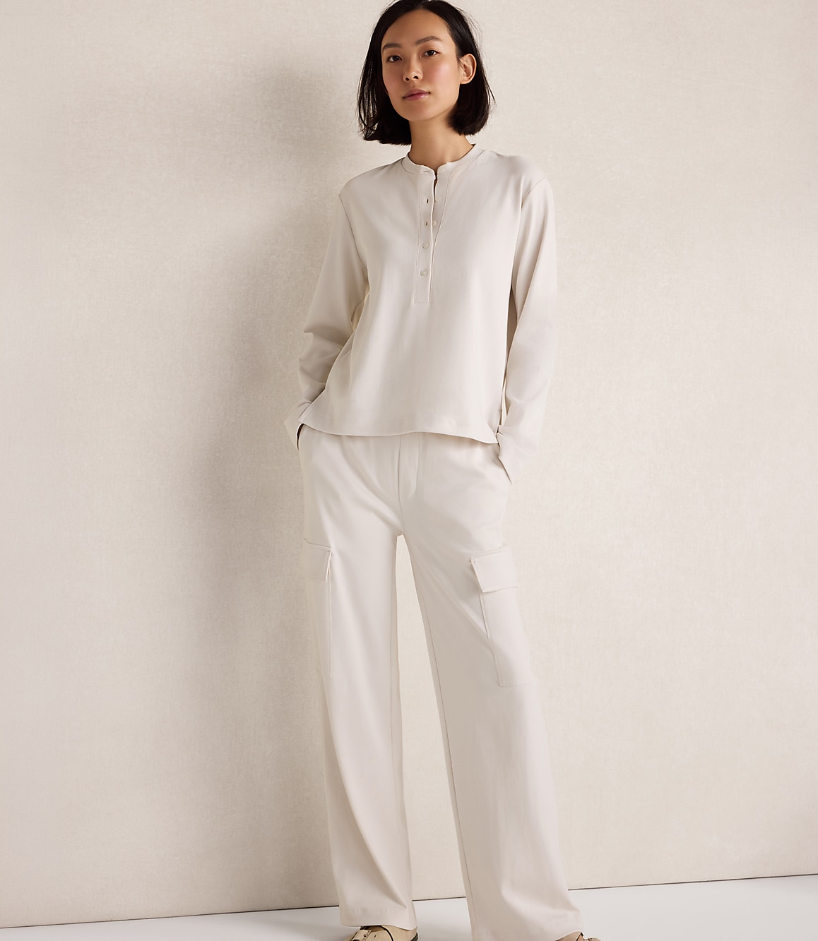 A woman in a cream-colored pajama set stands against a plain, textured wall.