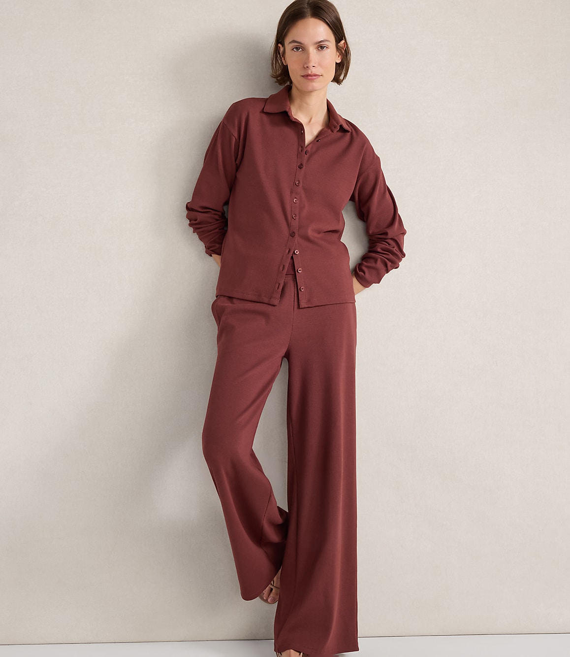 A woman in a maroon pajama set stands against a textured off-white wall.