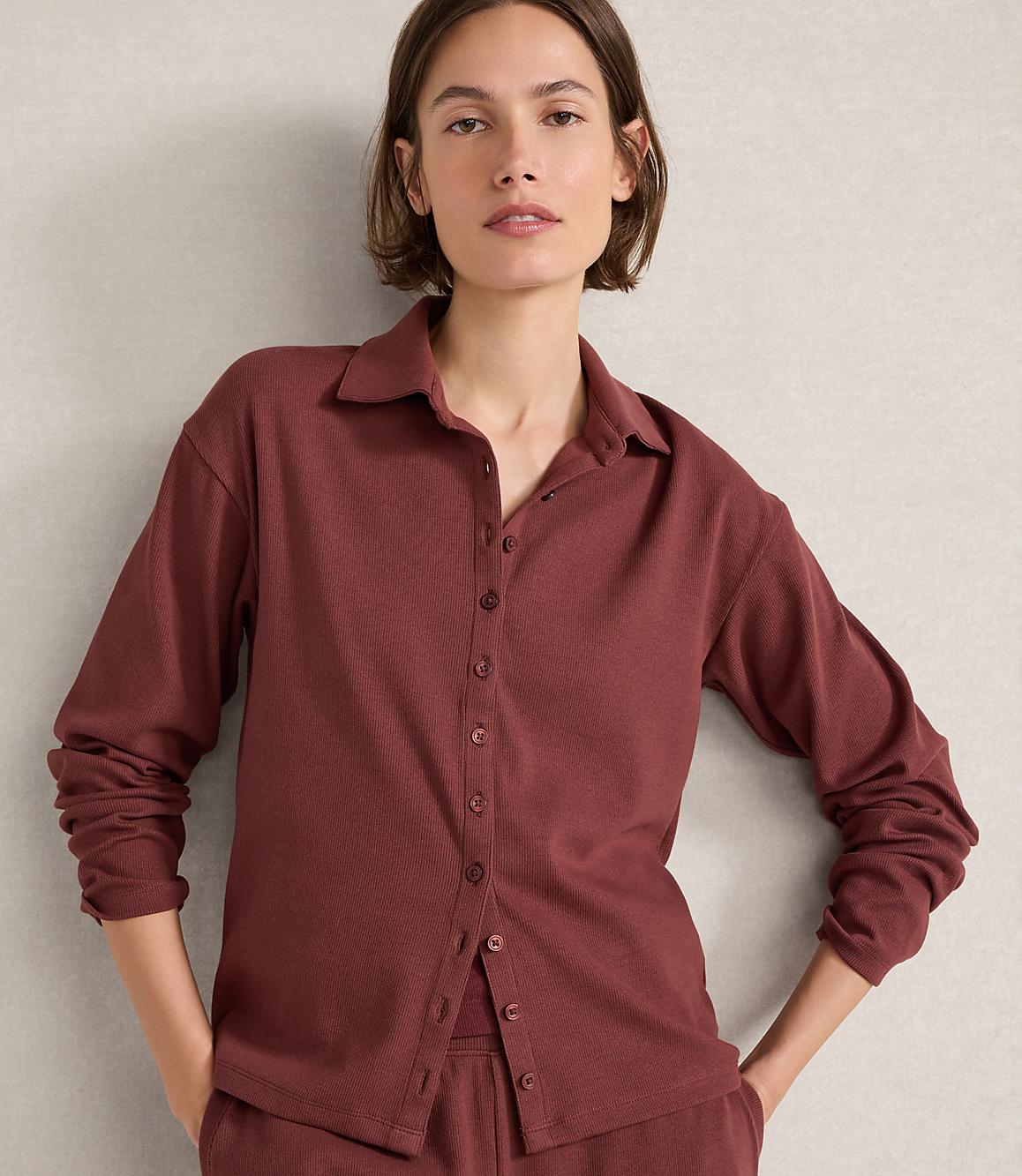 A woman in a maroon ribbed button-down shirt and matching pants stands against a textured gray wall.