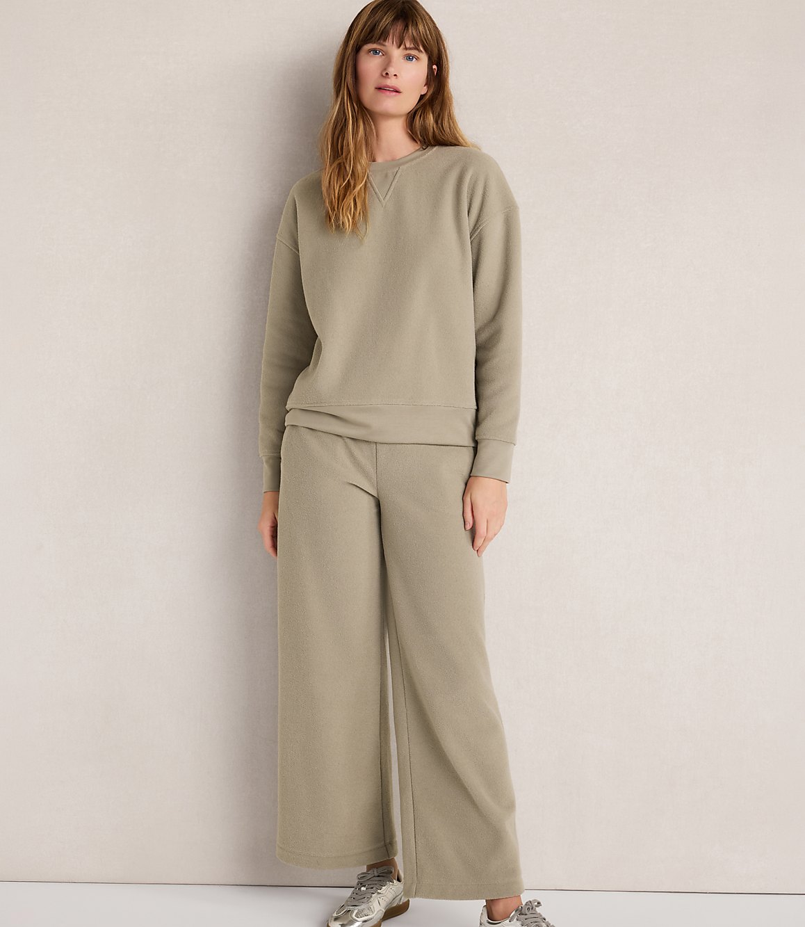 A woman in a light green sweatshirt and wide-leg pants stands against a plain, light-colored wall.