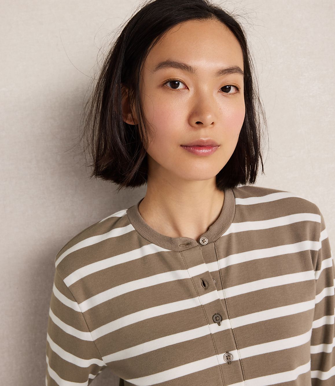 A young woman with short dark hair and a striped shirt is looking forward against a textured gray background.