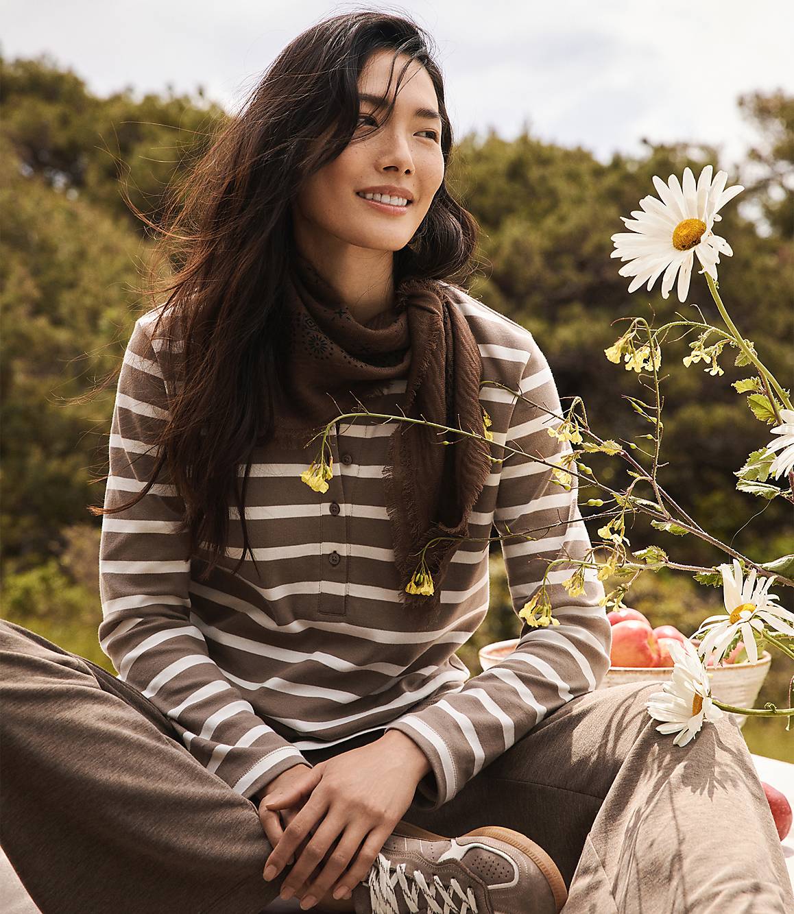 A woman in a striped shirt and brown pants sits outdoors with daisies and apples in the foreground, and a blurred green landscape in the background.