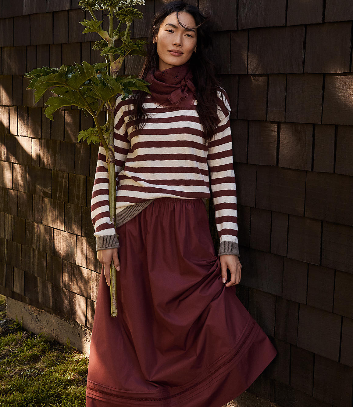 A woman in a striped sweater and maroon skirt stands in front of a wooden wall, holding a leafy green plant.