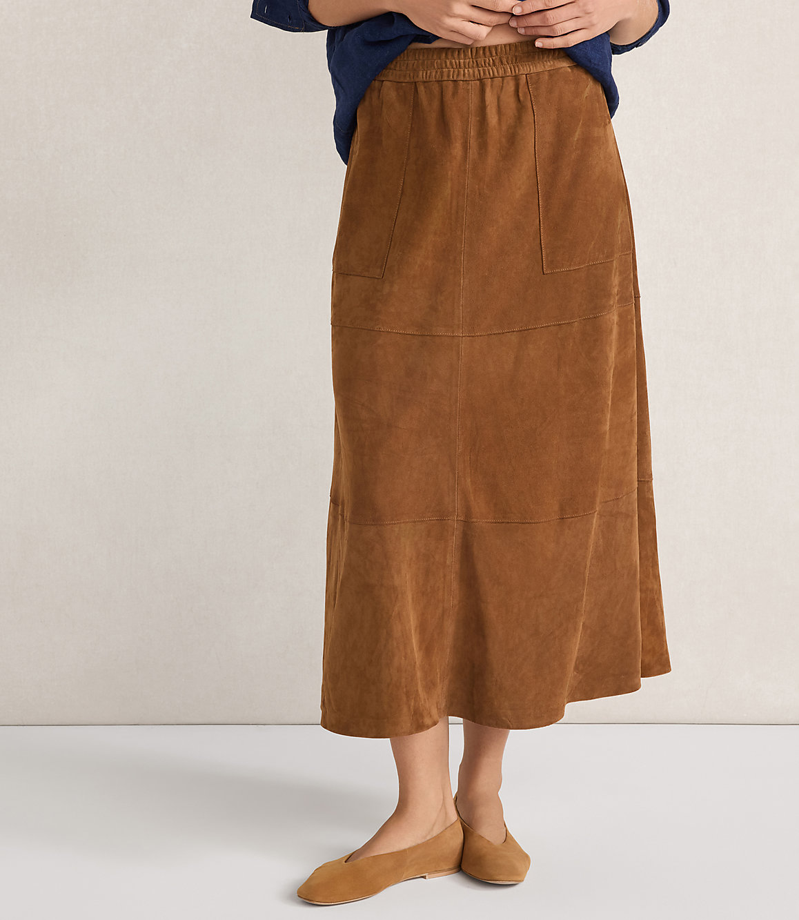 A person stands wearing a brown suede midi skirt and matching flats against a plain, light-colored background.