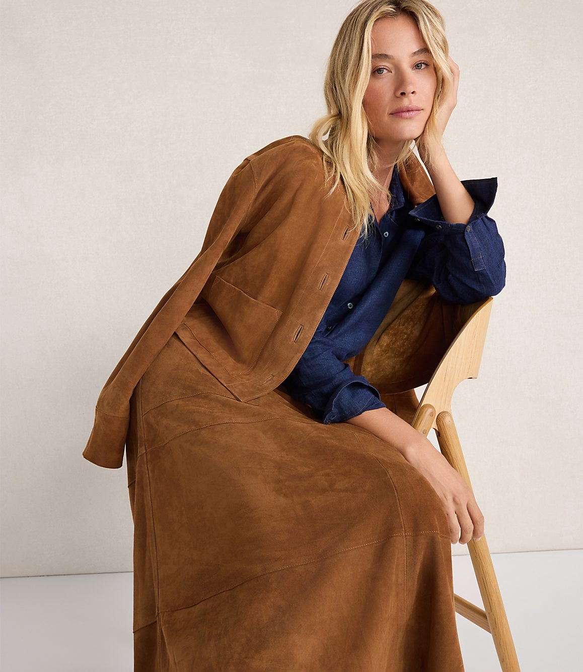 A blonde woman in a brown suede jacket and skirt sits on a wooden chair against a plain background.