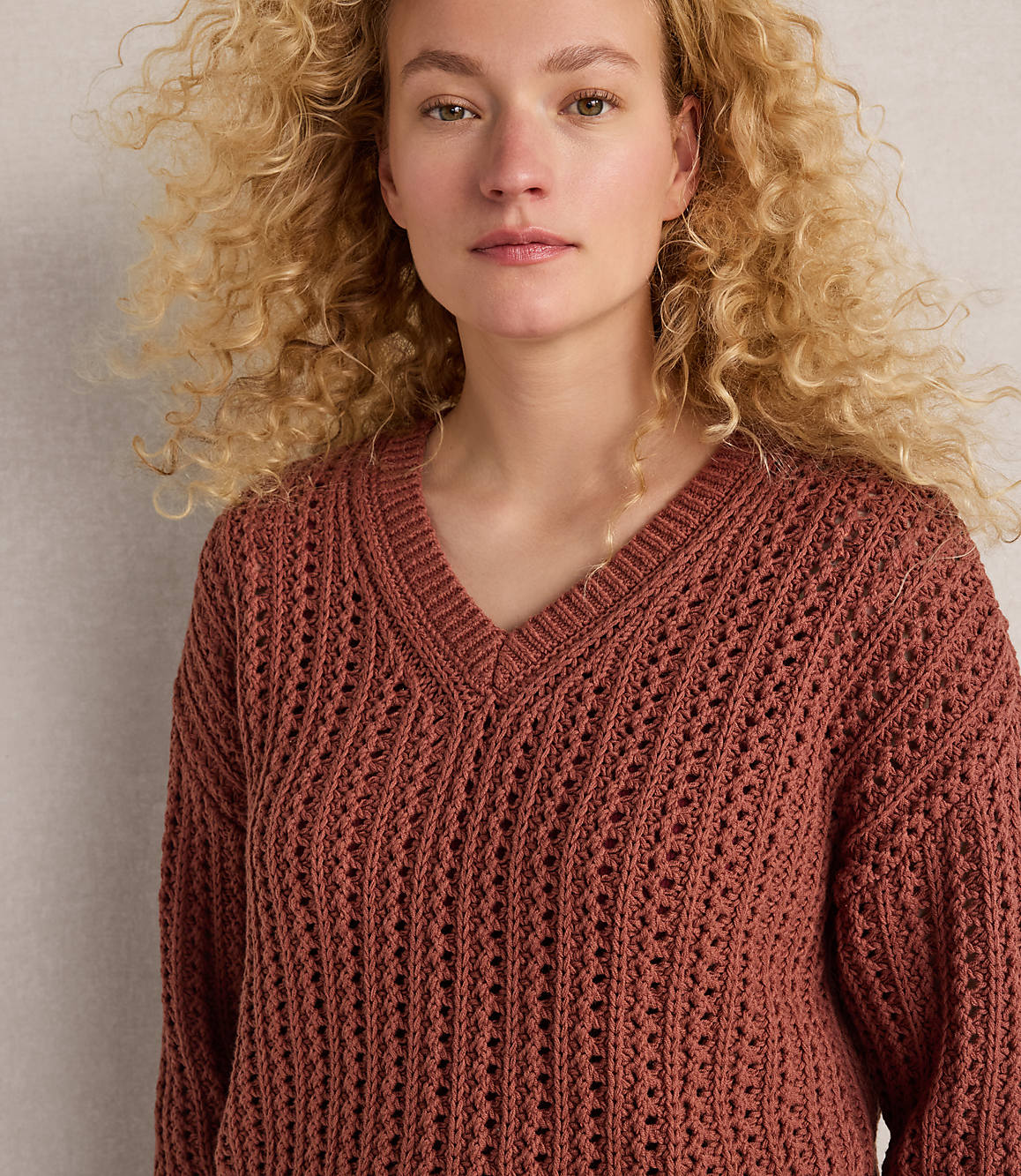 A young woman with curly blonde hair wears a rust-colored v-neck sweater in the foreground, against a plain, light-colored background.
