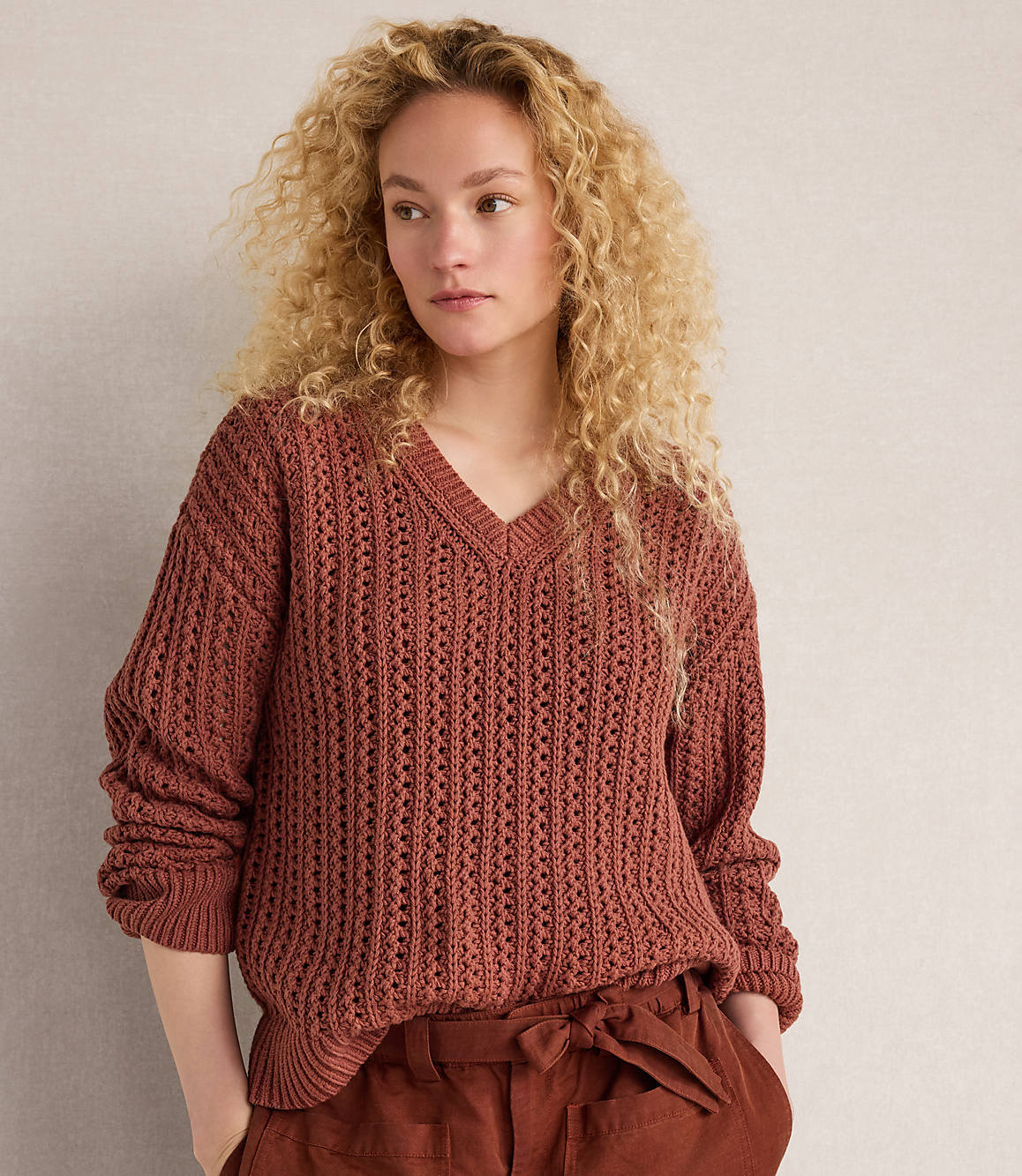 A young woman with curly blonde hair poses in a rust-colored, open-knit V-neck sweater and matching pants against a plain, light-colored wall.