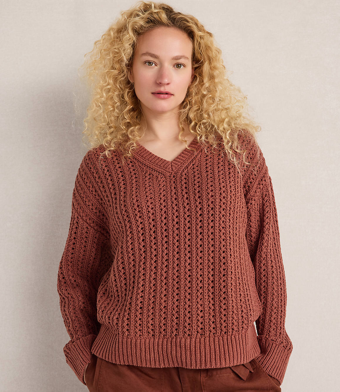 A woman with curly blonde hair wears a rust-colored v-neck sweater and brown pants against a plain, light-colored background.