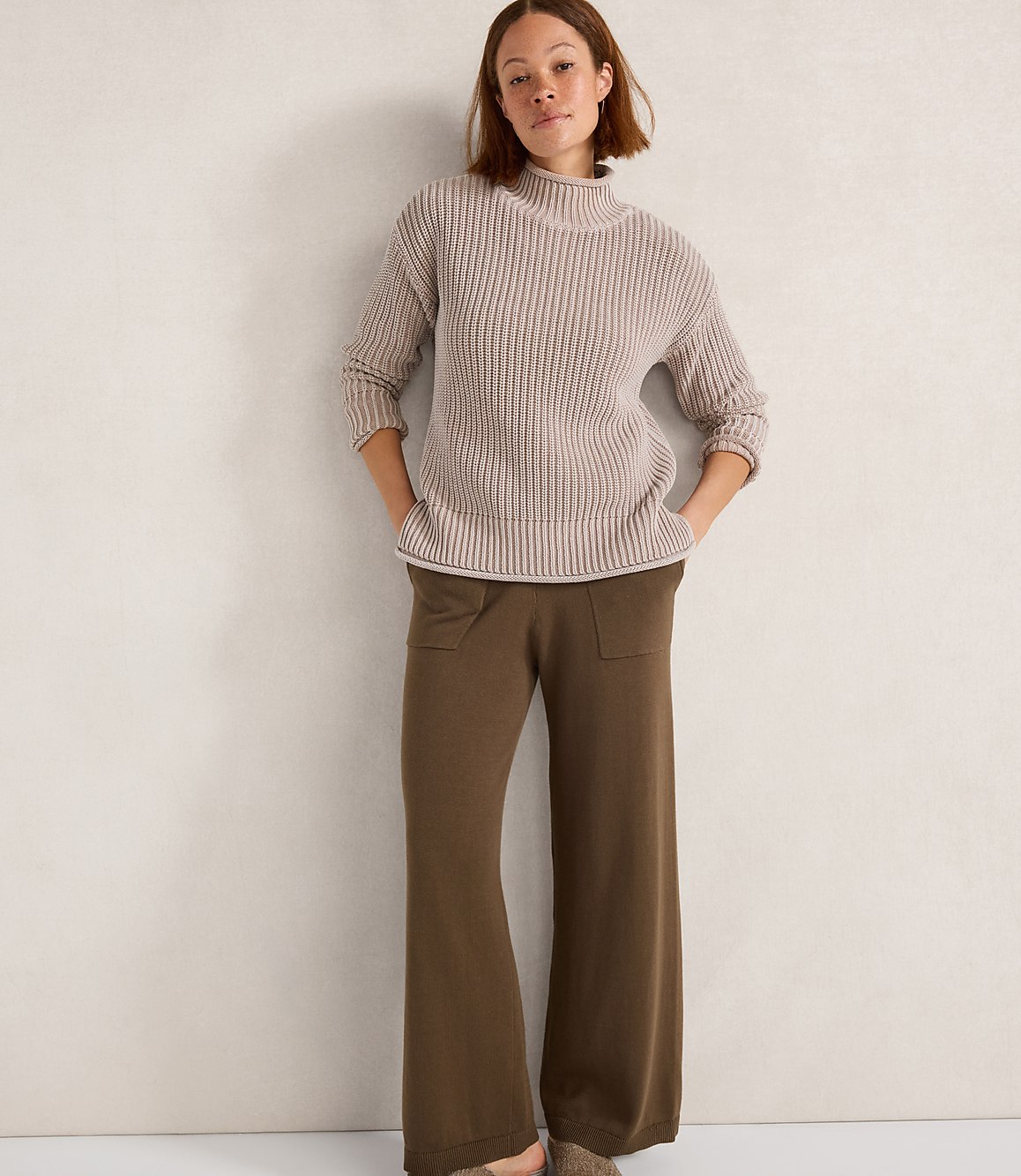 A woman in a beige ribbed turtleneck sweater and olive green wide-leg pants stands against a textured off-white wall.