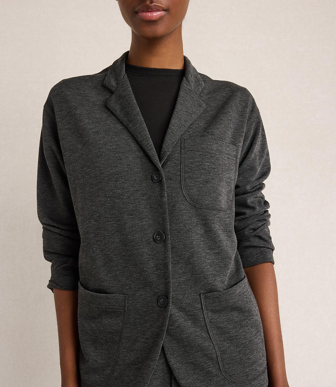 A woman in a dark grey blazer stands against a plain, light grey background.