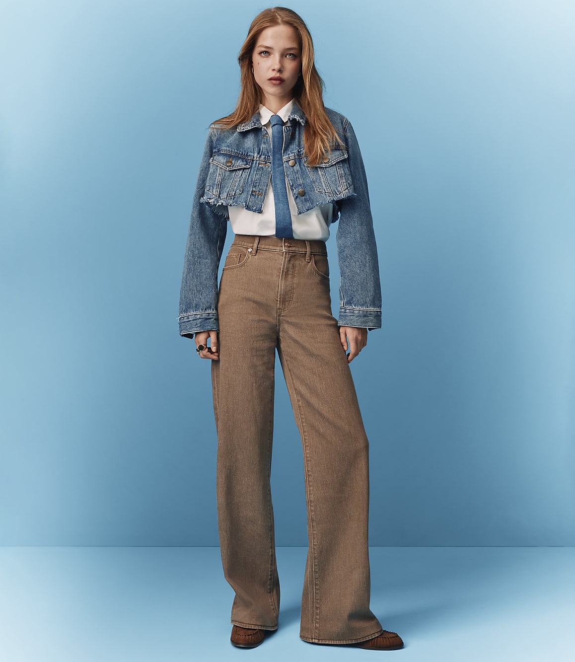 A young woman in a denim jacket and wide-leg pants stands against a solid blue background.