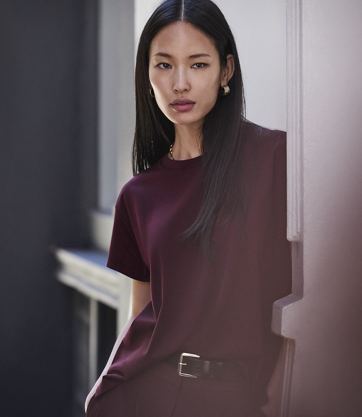 A young woman with long dark hair, wearing a maroon t-shirt and pants, stands in front of a blurred background of a building.