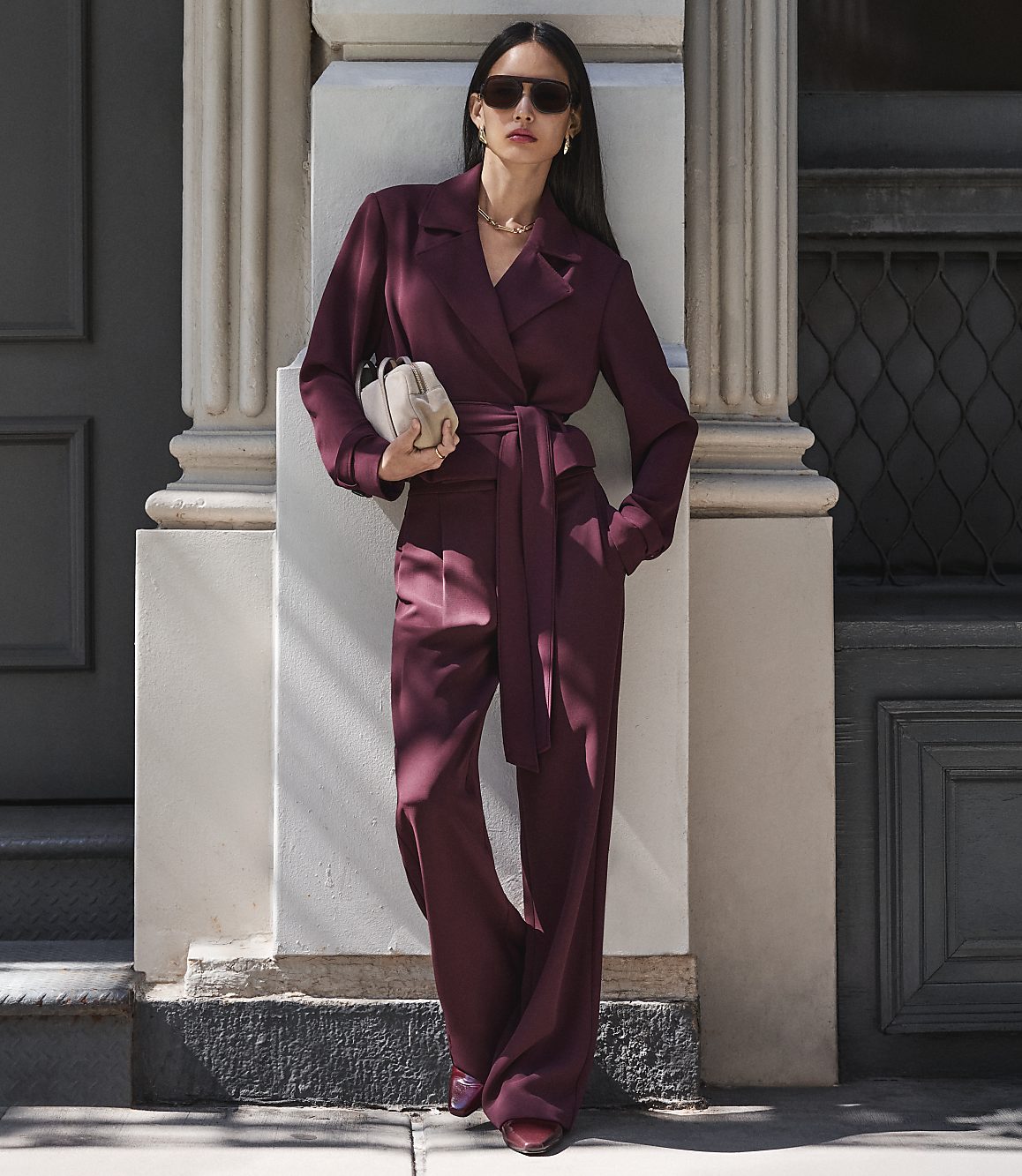 A woman in a burgundy jumpsuit stands against a building with a column and a dark door.