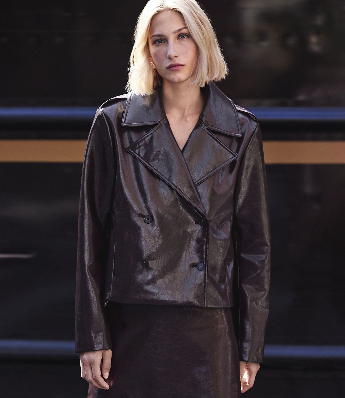 A blonde woman in a shiny brown jacket and skirt stands against a dark, textured background.