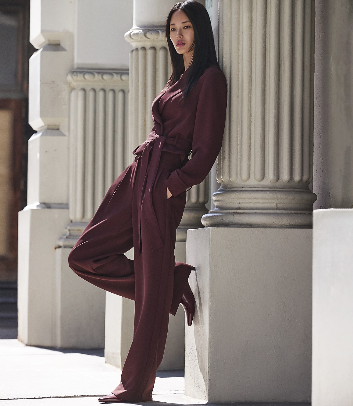 A woman in a maroon jumpsuit leans against a fluted column in front of a building.