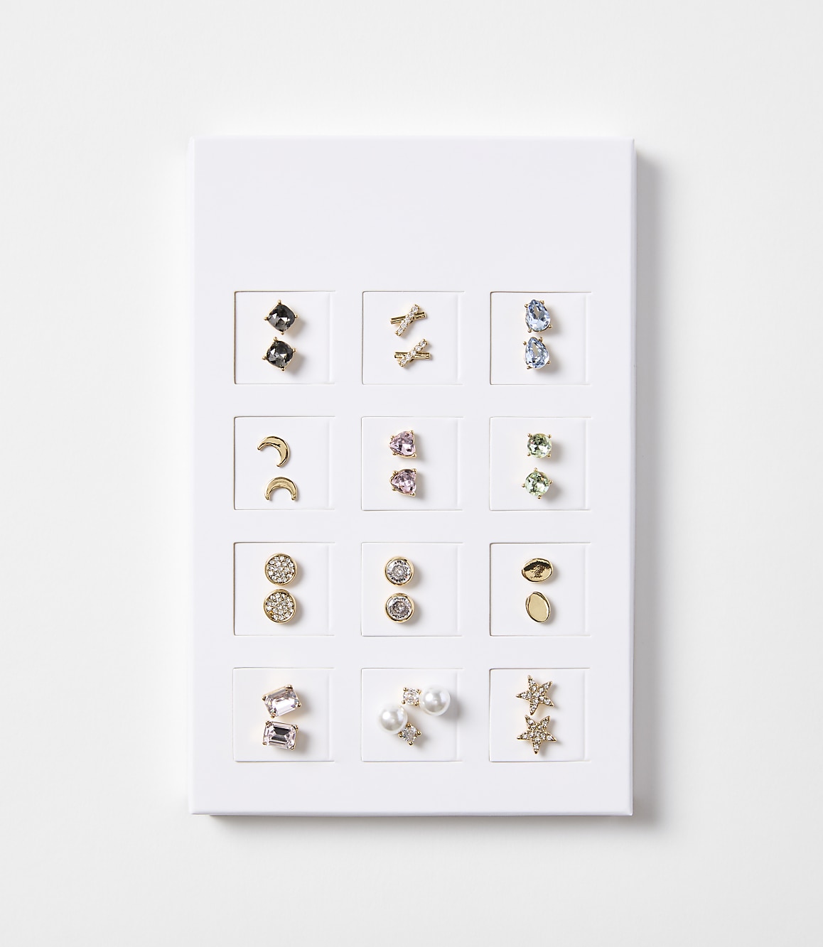 A white display board showcases twelve pairs of earrings, including studs, celestial shapes, and geometric designs, against a plain white background.