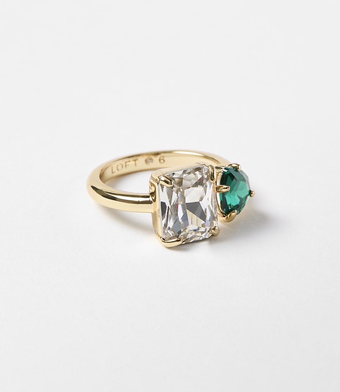 A gold ring with a rectangular clear stone and a heart-shaped green stone sits on a plain white background.