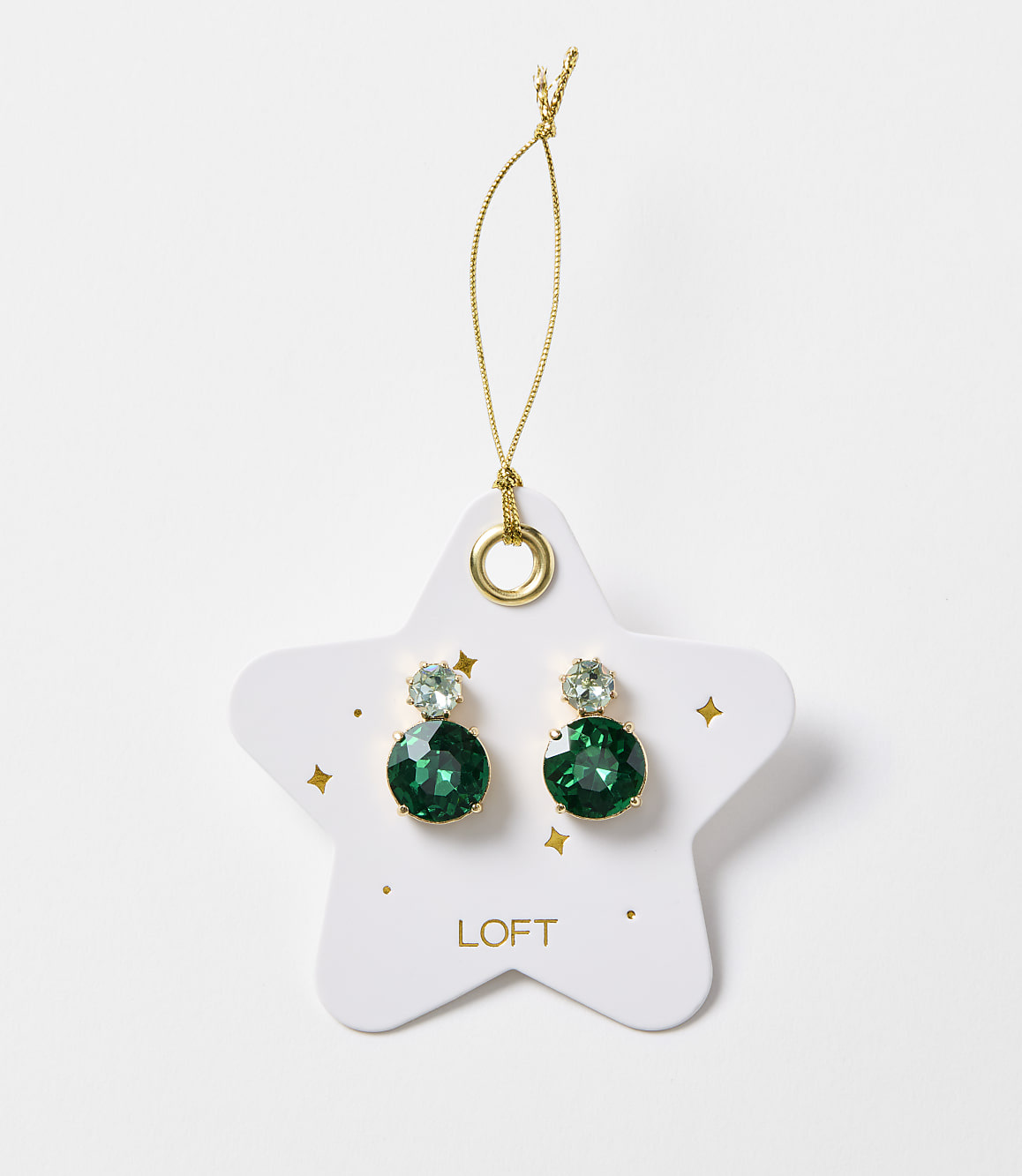 A pair of emerald green gemstone earrings dangle from a gold string attached to a white star-shaped card with gold accents and the "LOFT" logo, set against a plain white background.