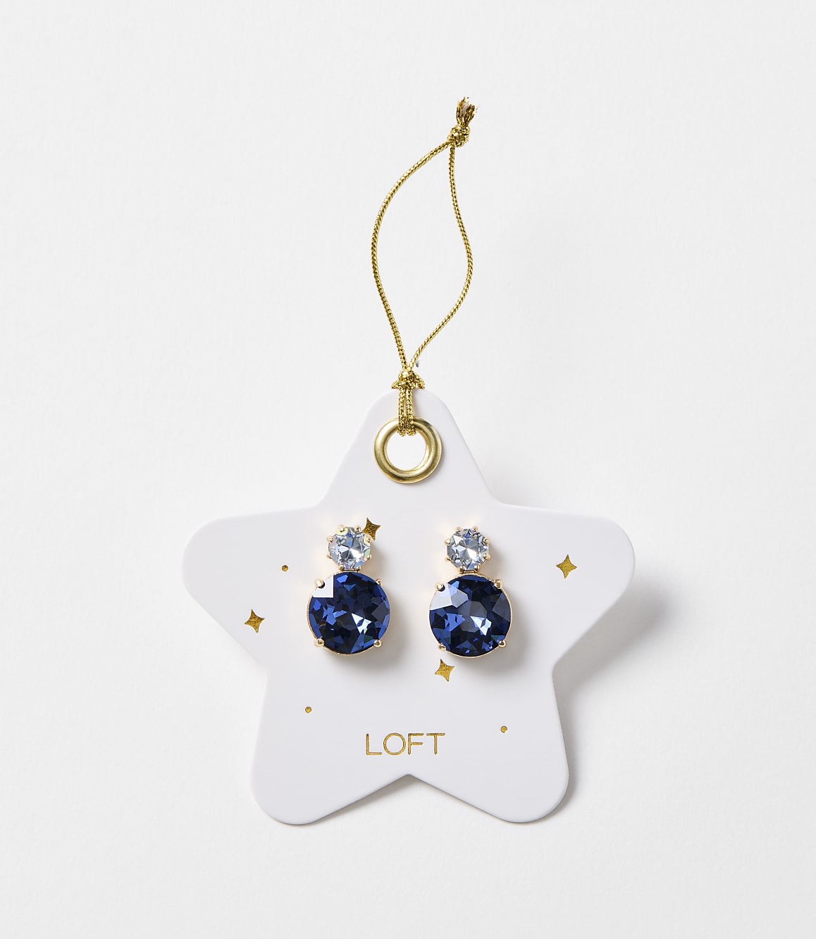 A pair of blue gem earrings are displayed on a white star-shaped card with gold accents and the "LOFT" logo, against a plain white background.