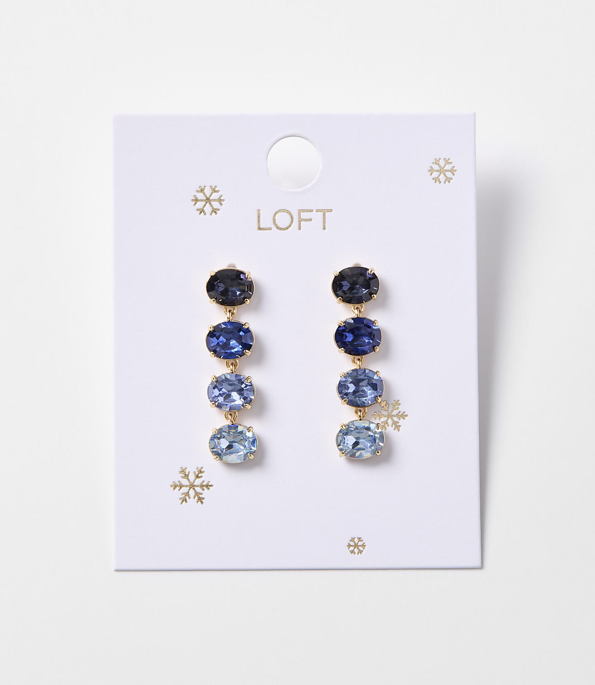 A pair of blue gemstone drop earrings with gold accents hangs from a white card with snowflake designs and the "LOFT" logo.