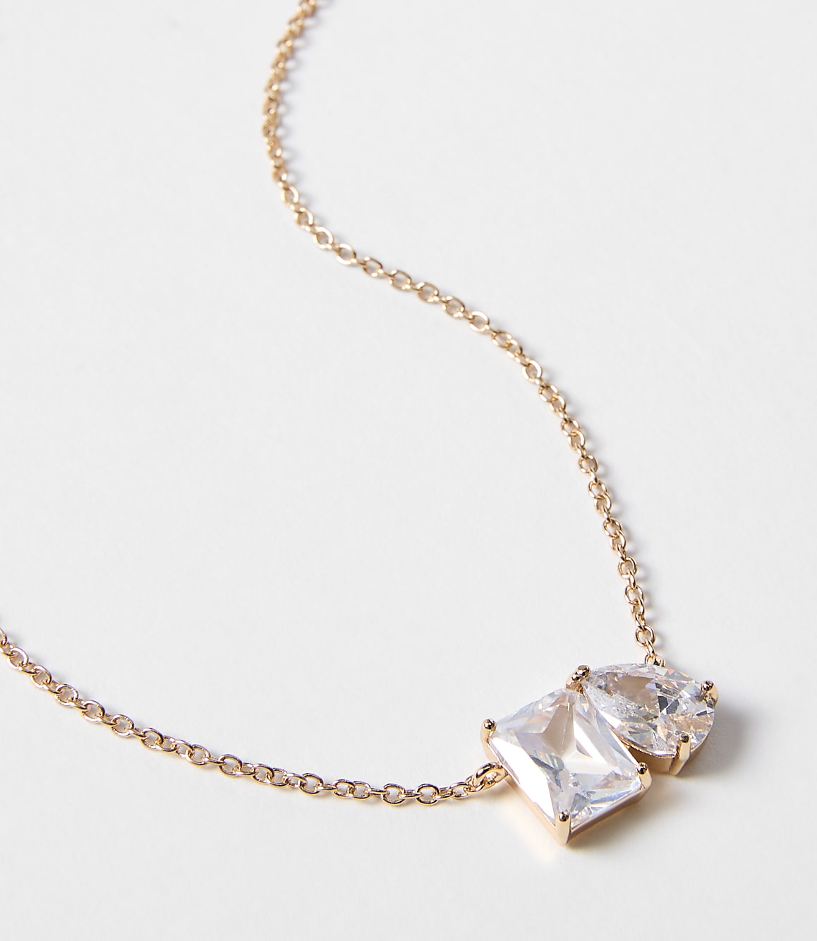 A delicate rose gold chain necklace with two clear, faceted gemstones, one rectangular and one pear-shaped, rests against a plain white background.
