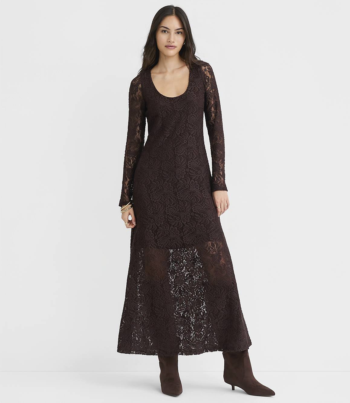 A woman in a long, dark brown lace dress and matching boots stands against a plain white background.