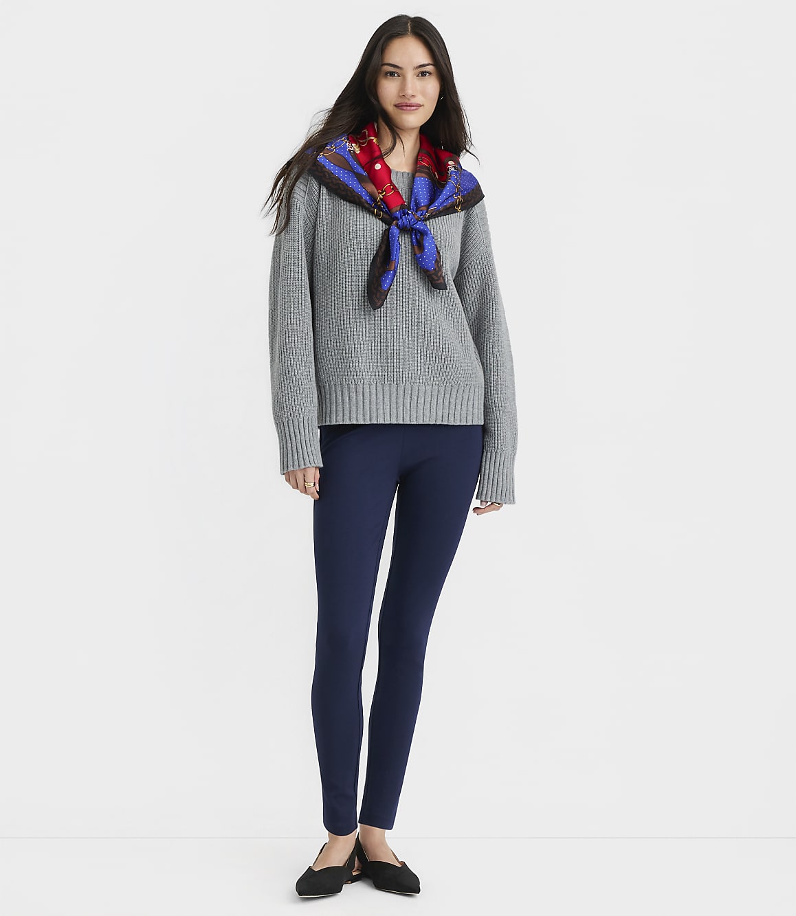 A woman in a gray sweater, navy leggings, and black flats stands against a plain gray background.