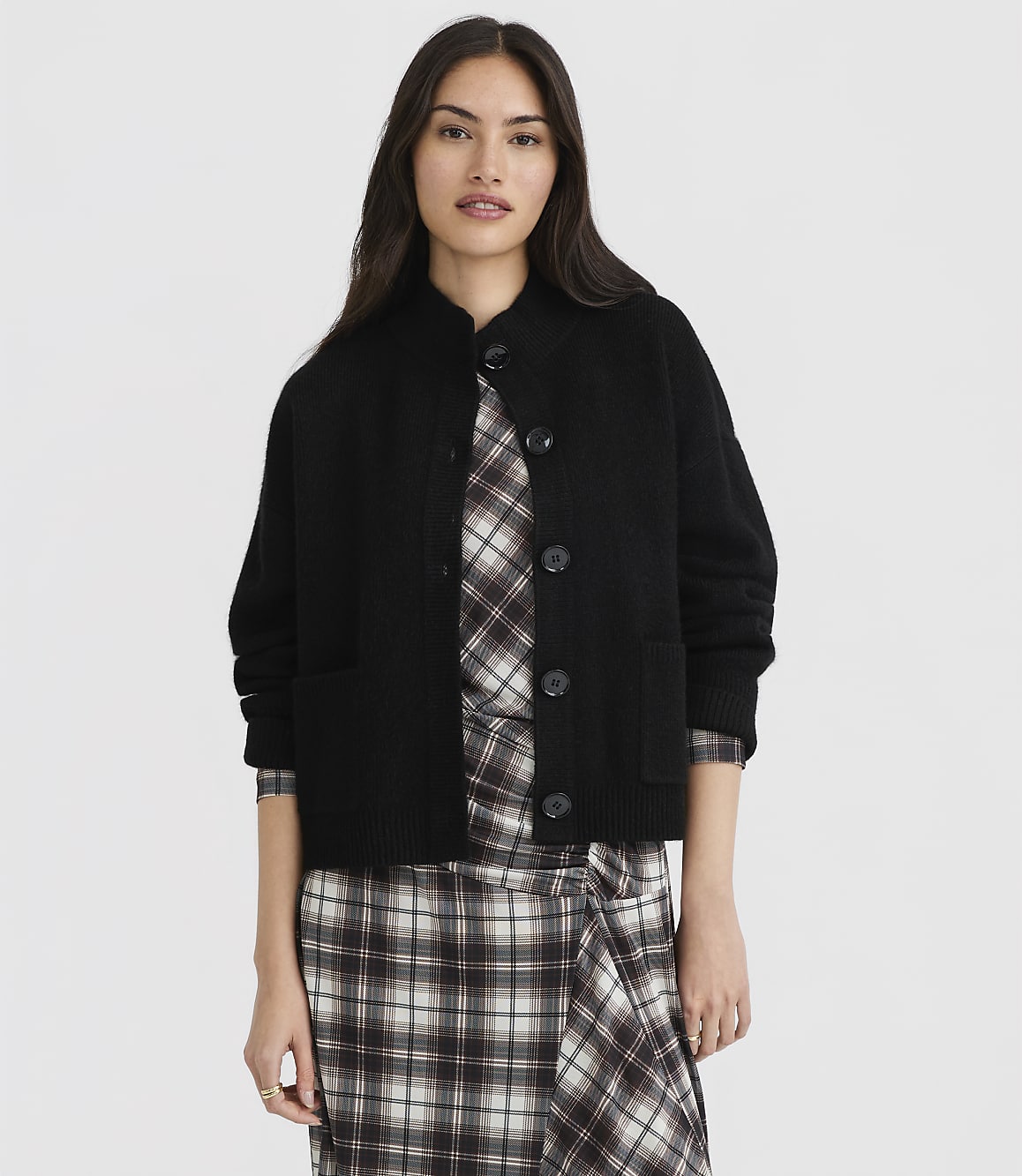A woman in a black cardigan and plaid dress stands against a plain white background.