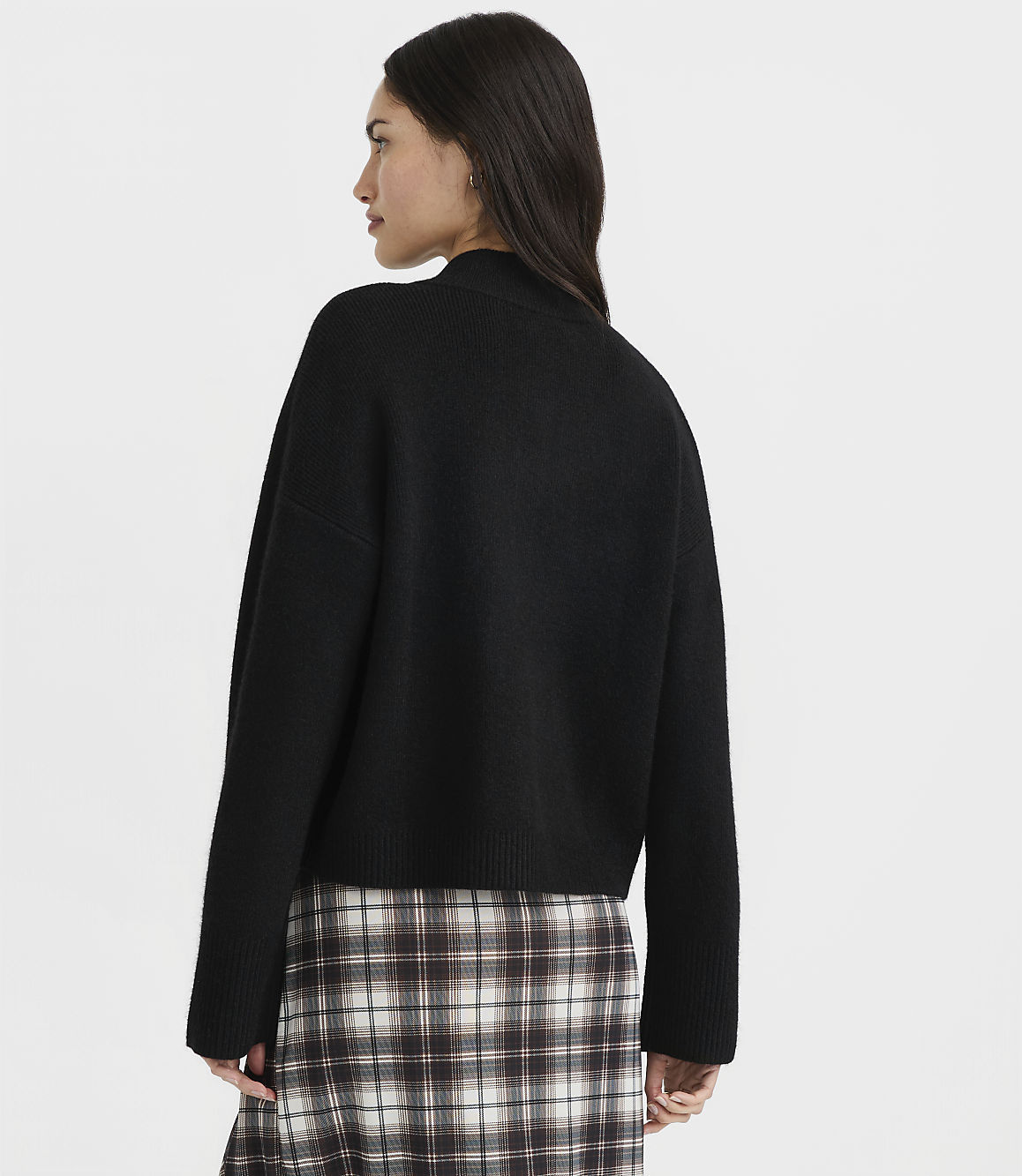 A woman in a black sweater and plaid skirt stands against a plain white background.