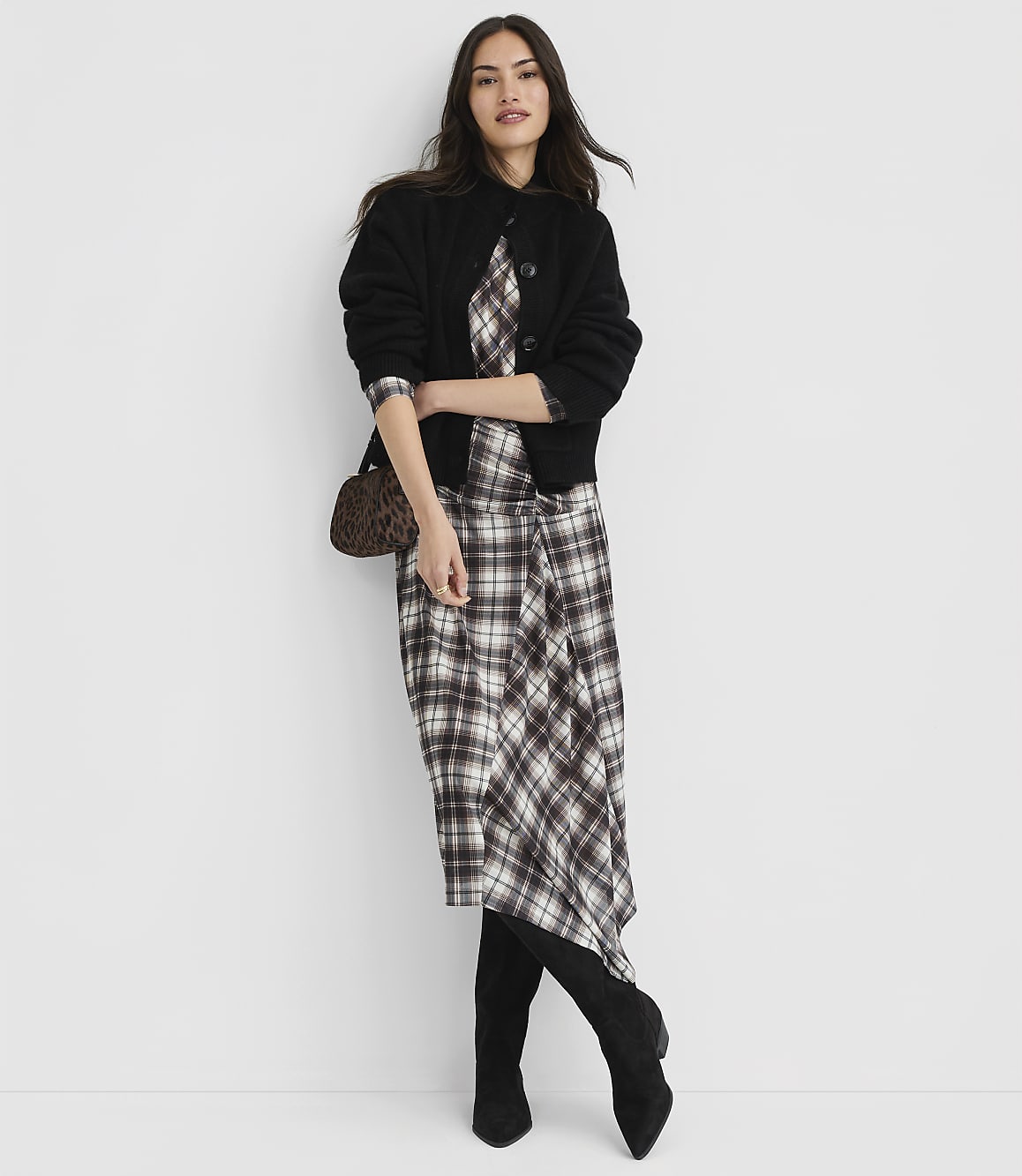 A woman in a plaid dress and black cardigan stands against a plain white background, accessorized with a leopard print clutch and black boots.