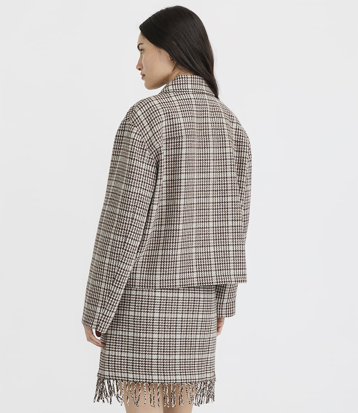 A woman in a plaid jacket and skirt with fringe stands against a plain white background.