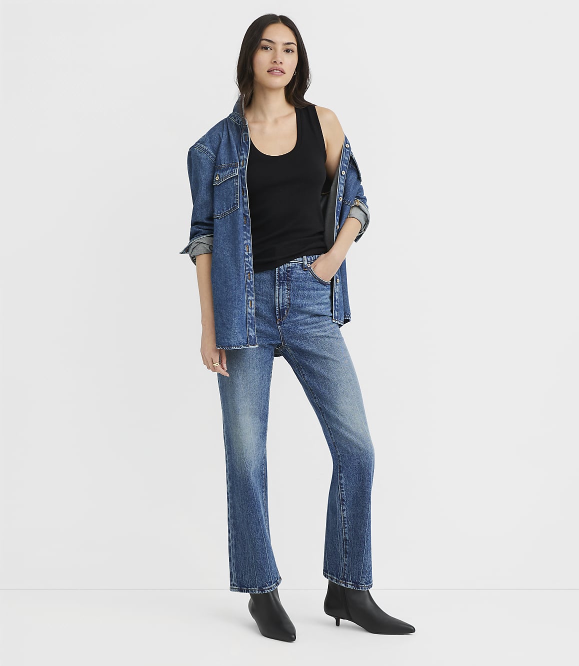 A woman in a black tank top, blue jeans, and a denim jacket stands against a plain white background.