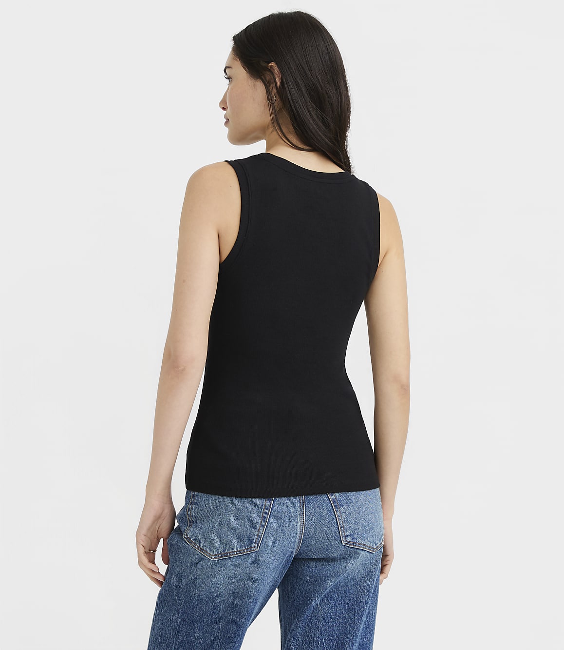 A woman in a black ribbed tank top and blue jeans stands against a plain white background.