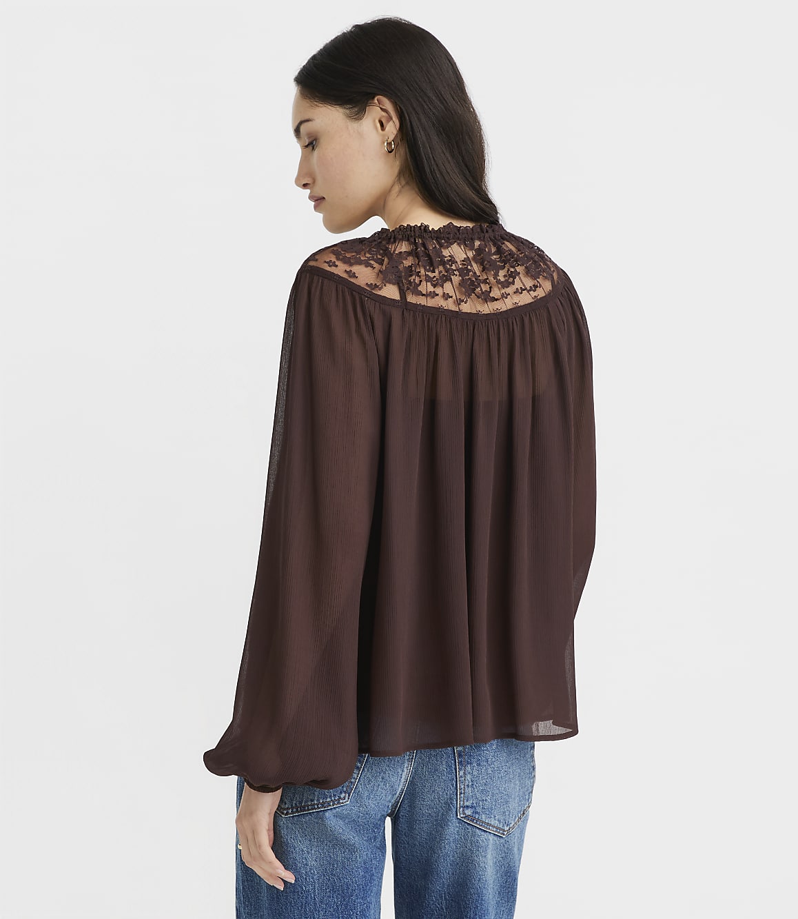 A woman in a brown lace-trimmed blouse and blue jeans stands against a plain white background.