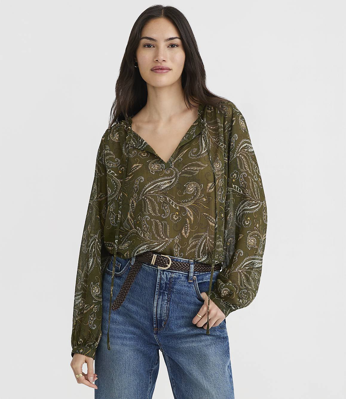 A woman in a green paisley blouse and blue jeans stands against a plain white background.
