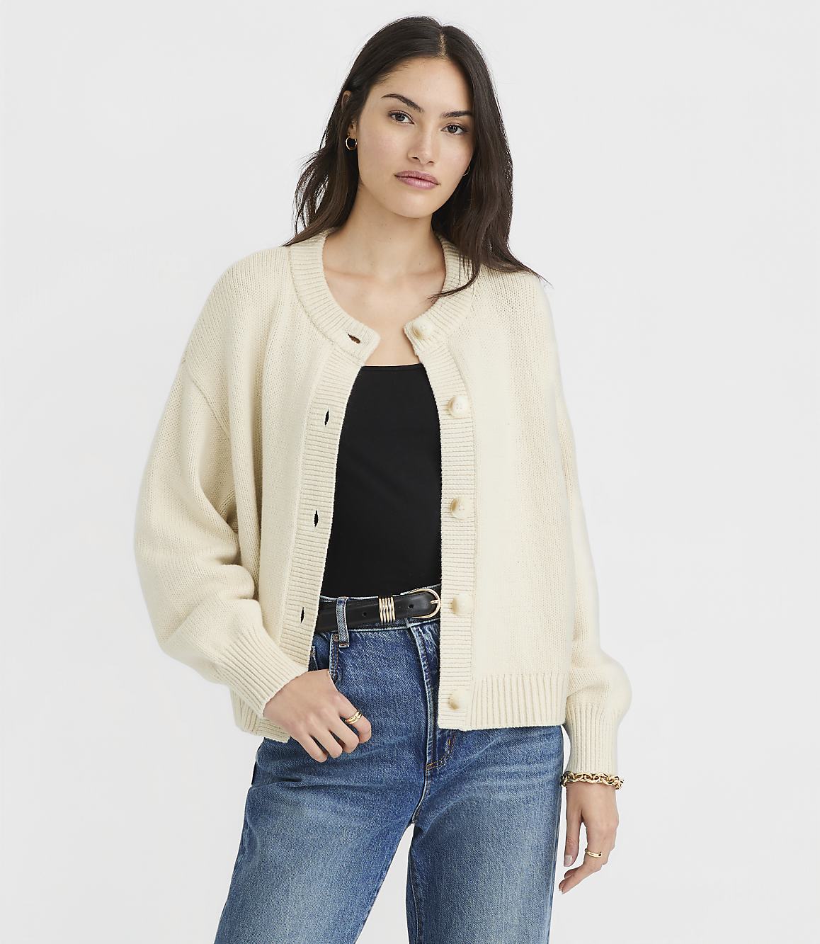 A woman in a cream cardigan and blue jeans stands against a plain white background.