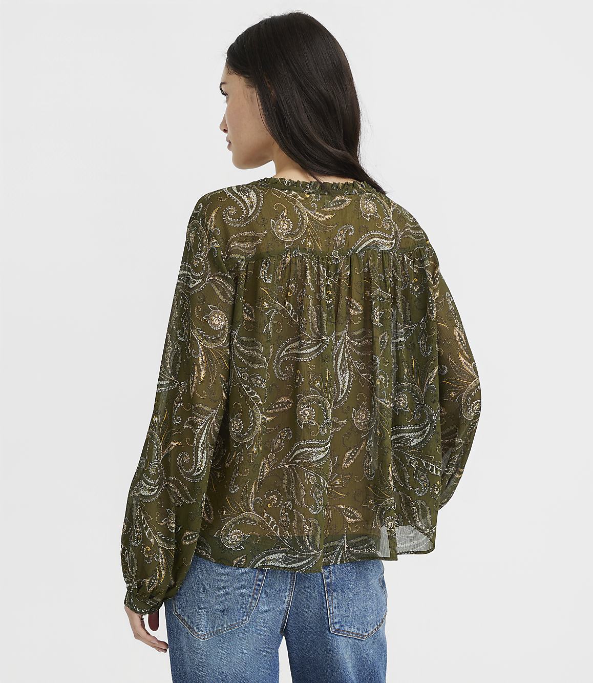 A woman in a green paisley blouse and blue jeans stands against a plain white background.