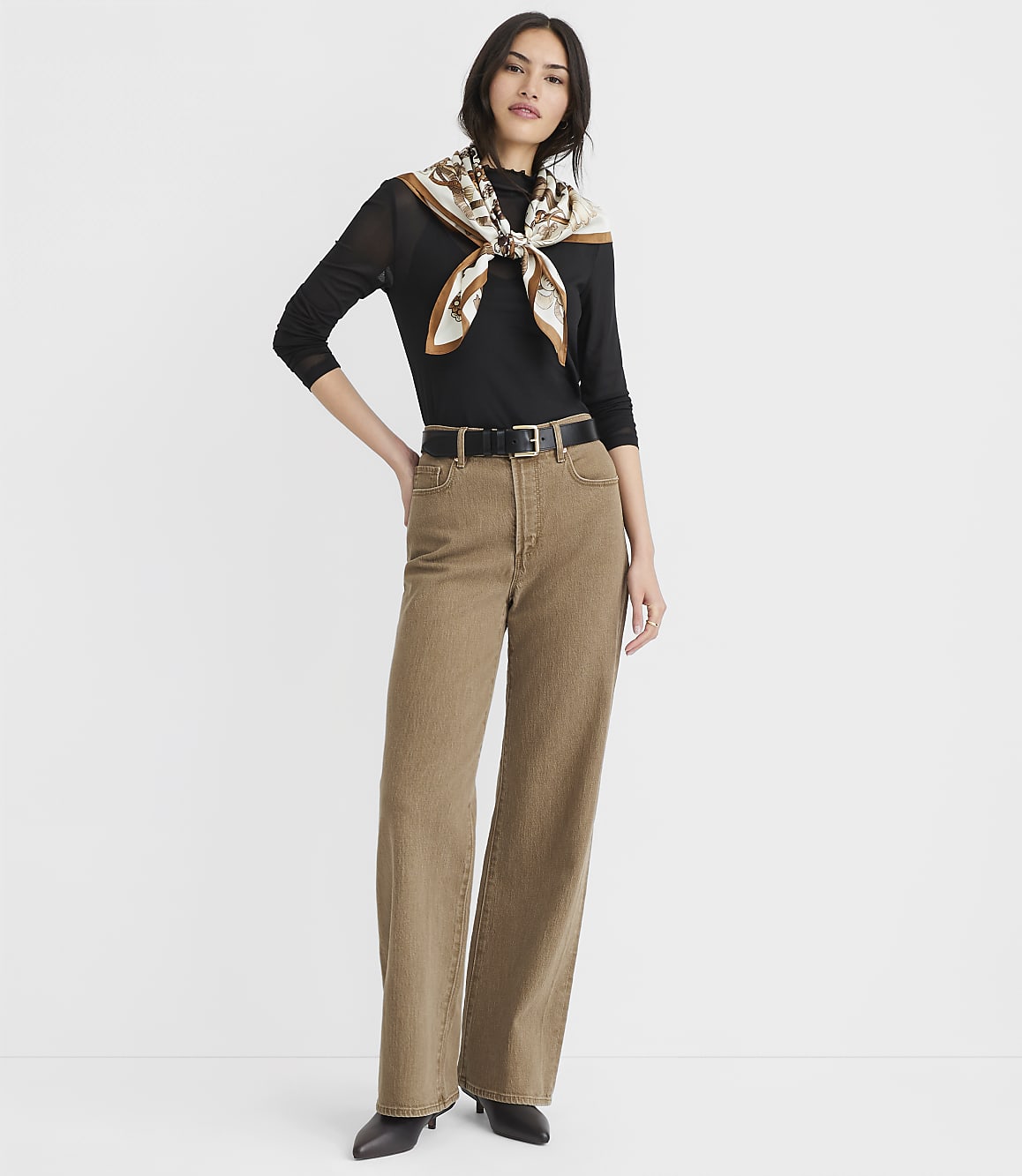 A woman in a black top, brown jeans, and a patterned scarf stands against a plain white background.
