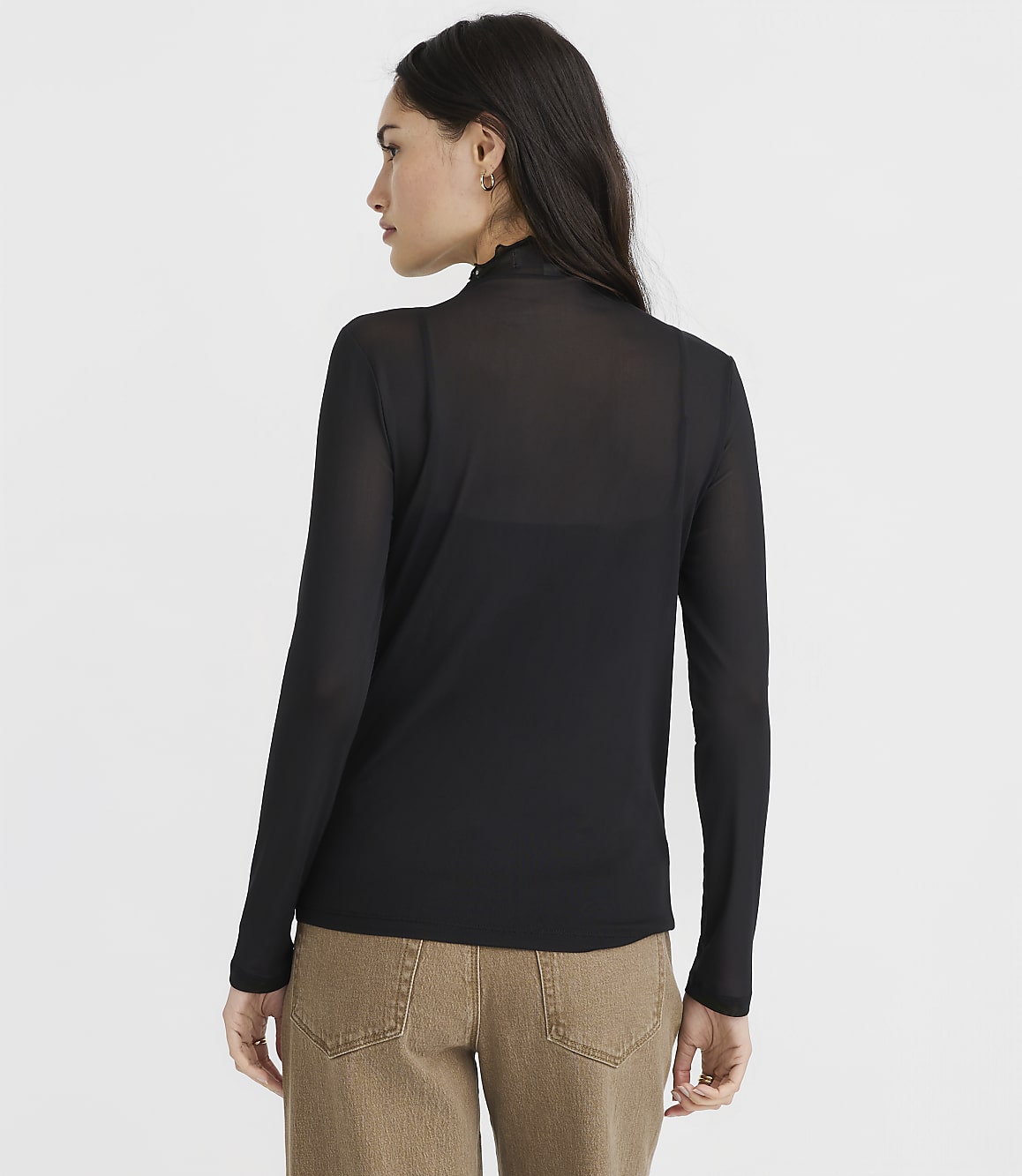A woman in a black sheer turtleneck and brown pants stands against a plain white background.