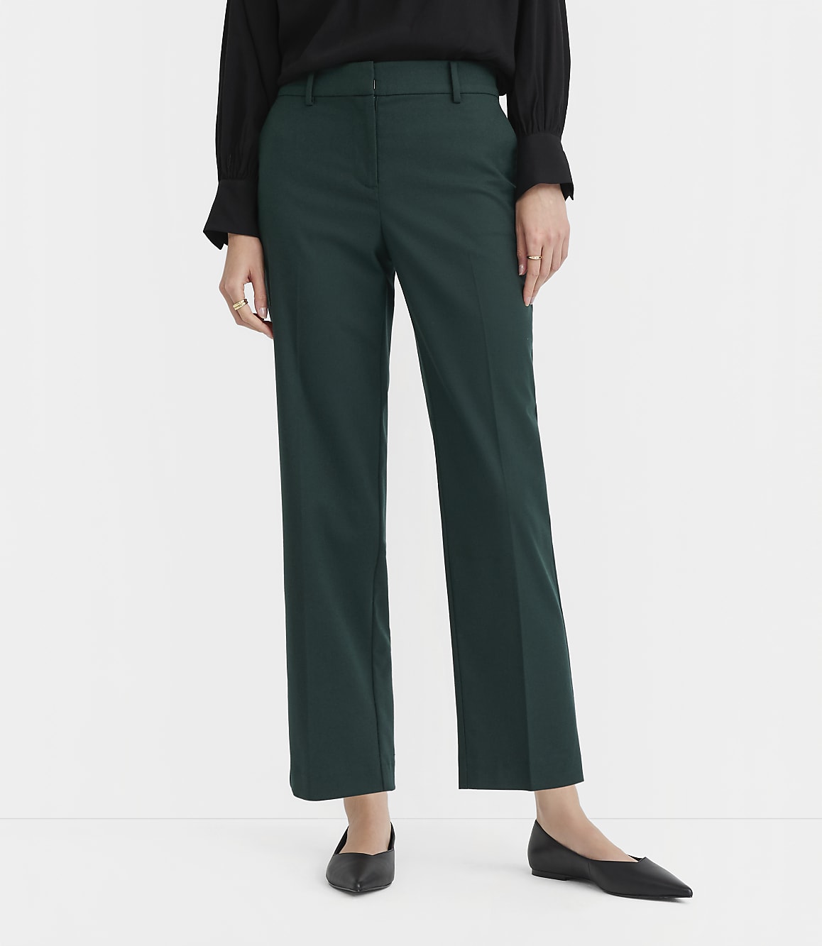 A person in a black top and dark green pants stands on a white floor against a white background.