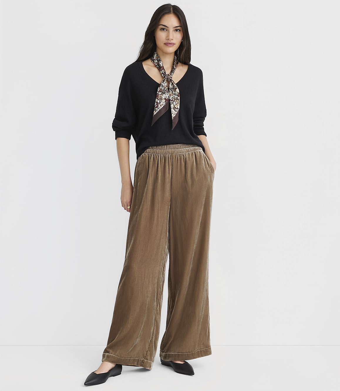 A woman in a black sweater and brown velvet pants stands against a plain white background.