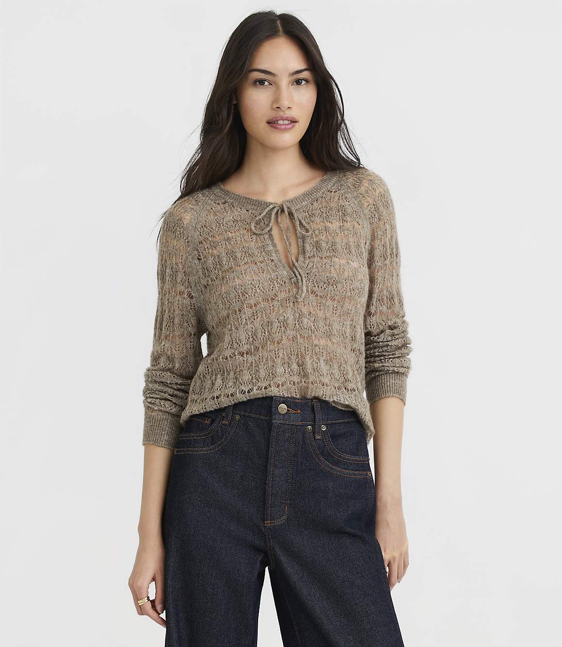 A woman in a light brown, open-knit sweater with a tie at the neck and dark wash jeans stands against a plain white background.