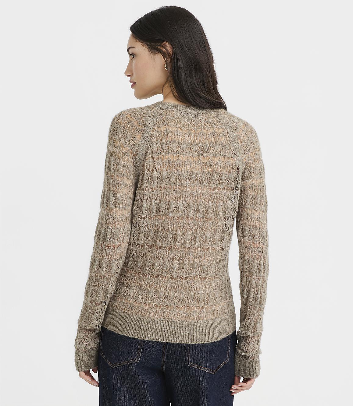 A woman in a sheer, textured brown sweater and dark jeans stands against a plain, light gray background.