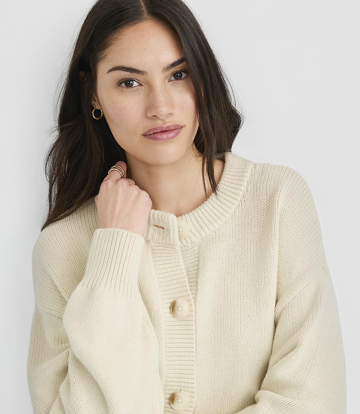 A woman in a cream-colored cardigan is captured in a close-up shot against a plain white background.