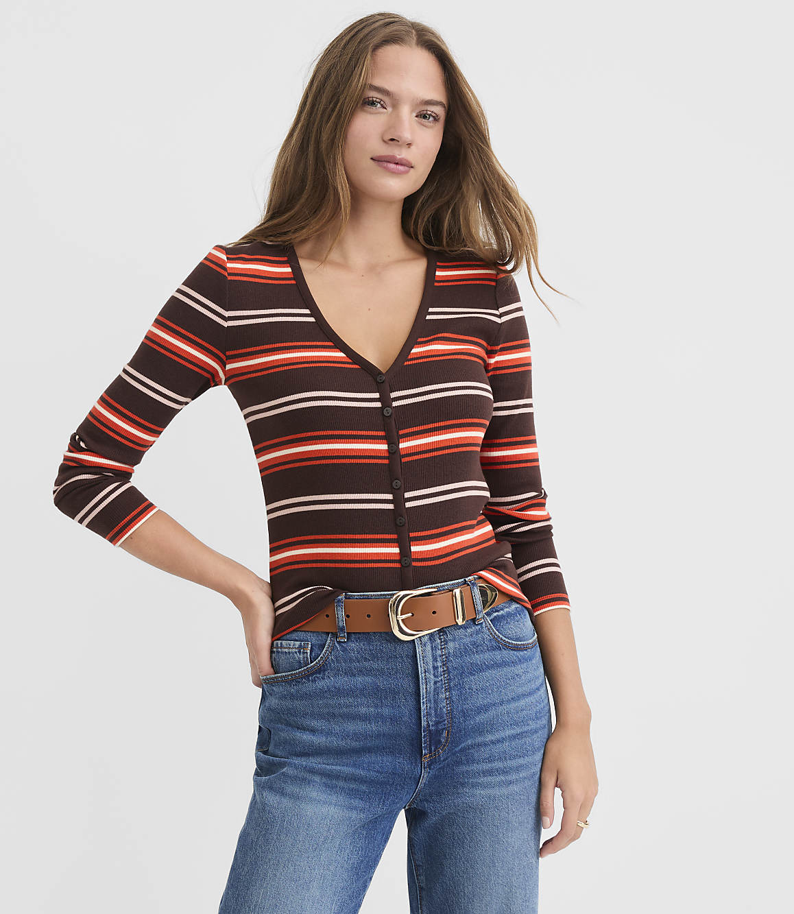 A woman in a striped v-neck sweater and jeans stands against a plain white background.