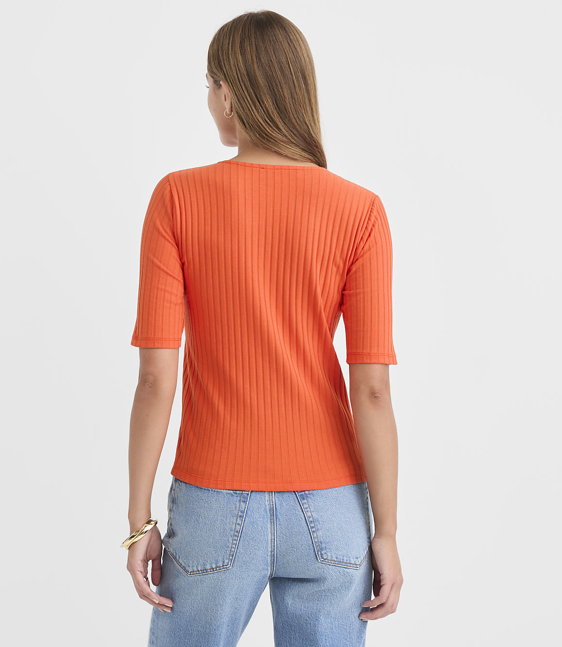 A woman in an orange ribbed t-shirt and blue jeans stands against a plain white background.