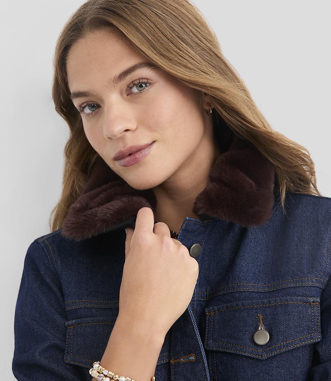 A young woman with brown hair and a denim jacket with a fur collar is shown against a plain gray background.