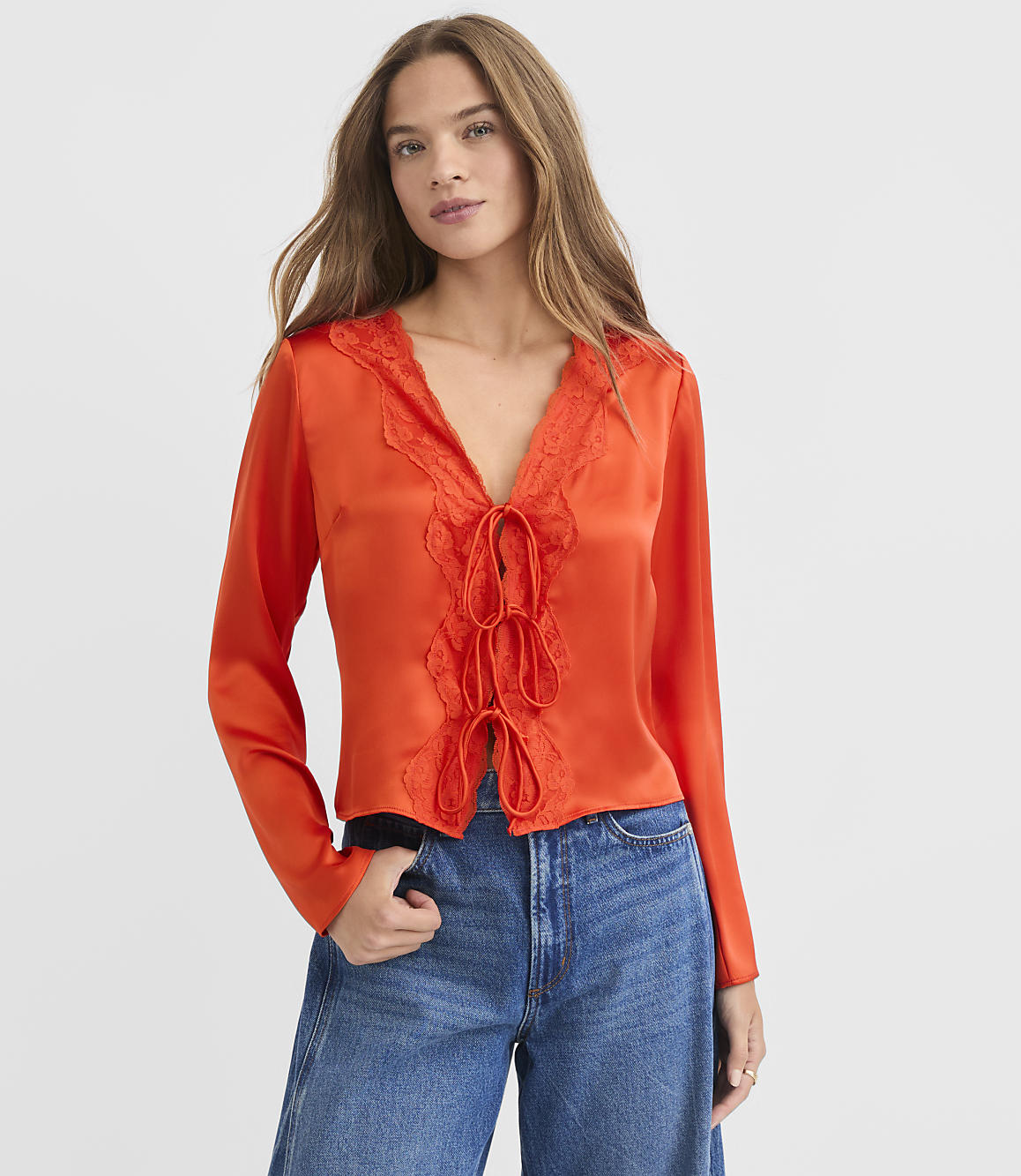 A woman in a vibrant orange lace-trimmed blouse and blue jeans stands against a plain white background.
