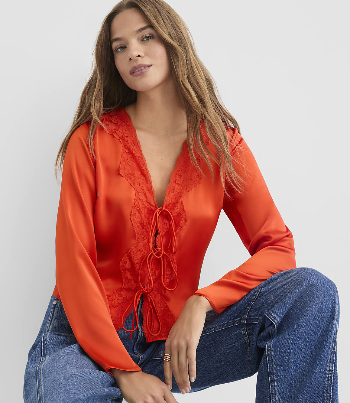 A woman in a vibrant orange lace-trimmed blouse and blue jeans poses against a plain white background.